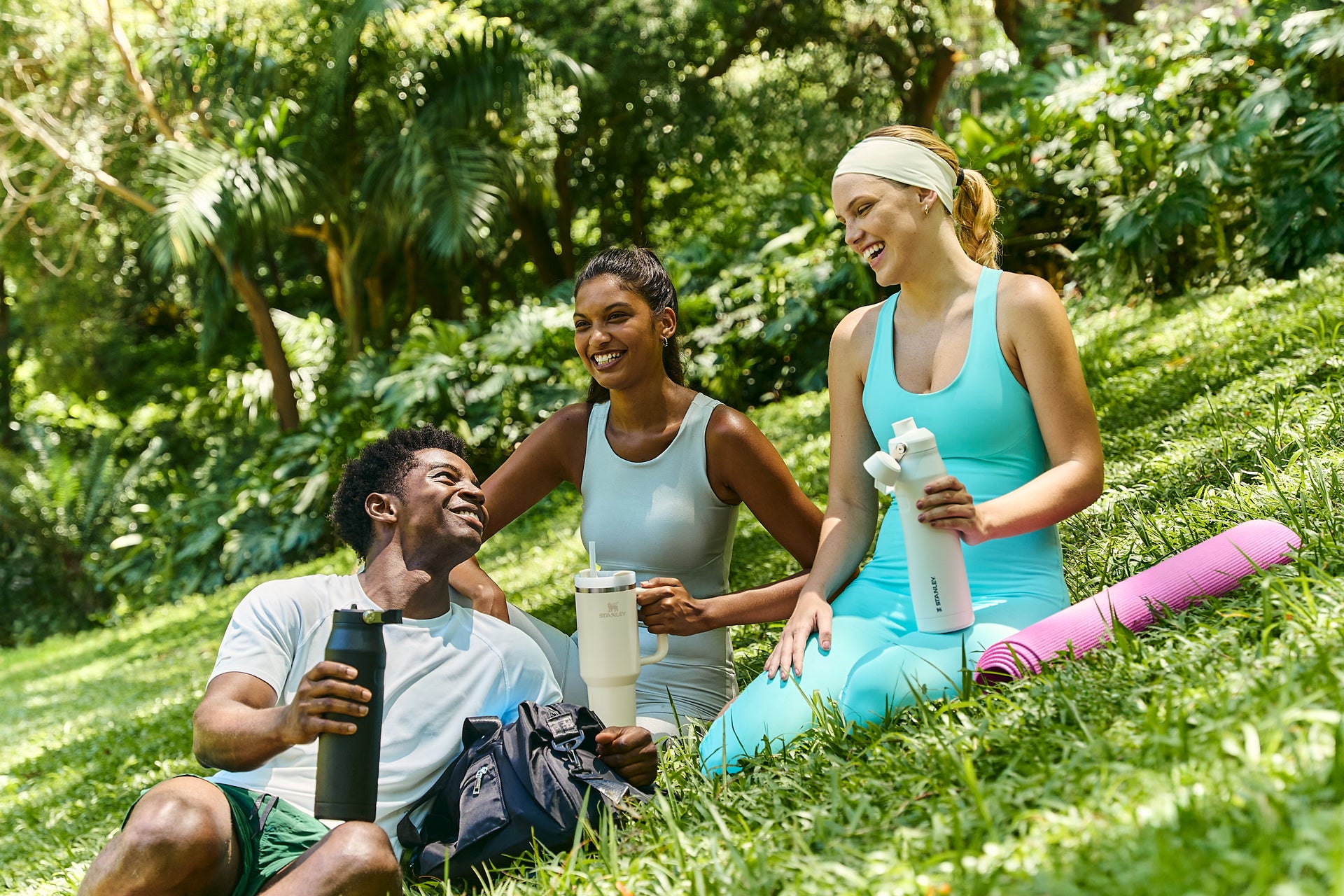 Três pessoas sorrindo sentadas em um gramado verde com garrafas térmicas Stanley. Ambiente ao ar livre com vegetação exuberante. Roupas esportivas e tapete de yoga ao lado. Ideal para atividades físicas e hidratação.