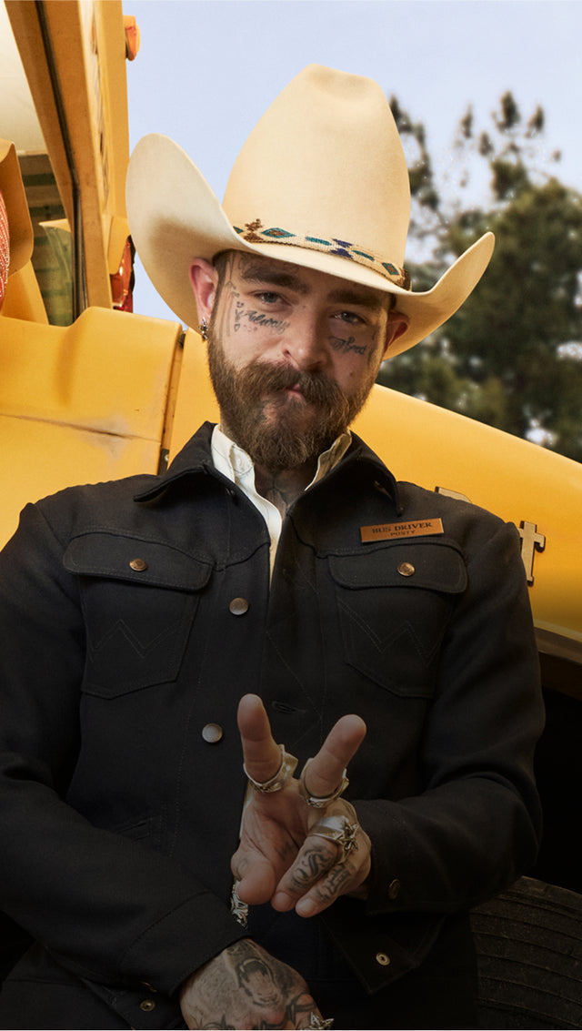 Homem com chapéu de cowboy. Ele tem barba e bigode, e seu rosto possui várias tatuagens. Está vestindo uma jaqueta preta com botões metálicos e uma camisa branca por baixo. Na jaqueta, há uma plaqueta com o nome "Bud Dwyer". Ele está fazendo um sinal de paz com os dedos. Ao fundo, há uma máquina amarela e algumas árvores desfocadas.