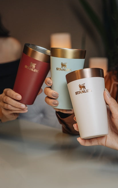 Três copos térmicos da marca Stanley estão sendo segurados por mãos diferentes, como se as pessoas estivessem brindando. Os copos têm um design elegante com uma faixa dourada na parte superior. Um copo é vermelho, outro é verde claro e o terceiro é branco. O logotipo da Stanley está impresso em dourado em cada copo.