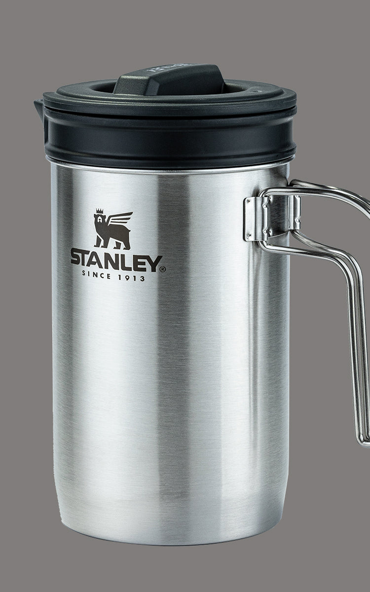 Copo térmico de aço inoxidável da marca Stanley. O copo é prateado com uma tampa preta e possui uma alça de metal dobrável. No corpo do copo, está o logotipo da Stanley, que inclui a imagem de um urso e a inscrição "STANLEY SINCE 1913". A tampa parece ser de plástico e tem um design que sugere que pode ser aberta ou fechada para beber.