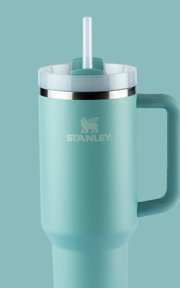 Copo térmico da marca Stanley em azul claro. Ele possui uma alça da mesma cor. A tampa é branca com um anel prateado e tem um canudo branco saindo do centro. O logotipo da Stanley, que inclui um urso alado, está impresso na frente do copo, logo abaixo da borda. O fundo da imagem é da mesma cor azul clara do copo, criando um efeito de continuidade.