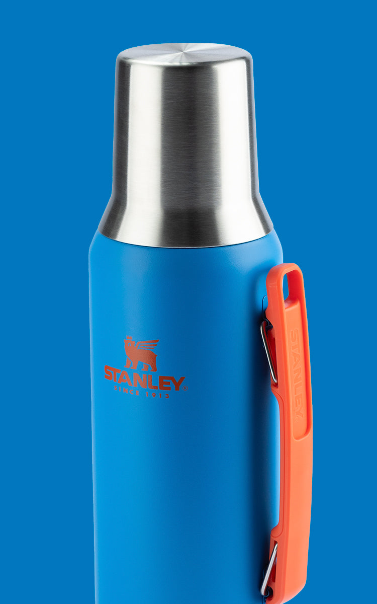 Garrafa térmica azul com tampa de aço inoxidável. A garrafa possui uma alça laranja na lateral e o logotipo da marca "Stanley" em laranja, com a imagem de um urso acima do nome. O fundo da imagem é azul.