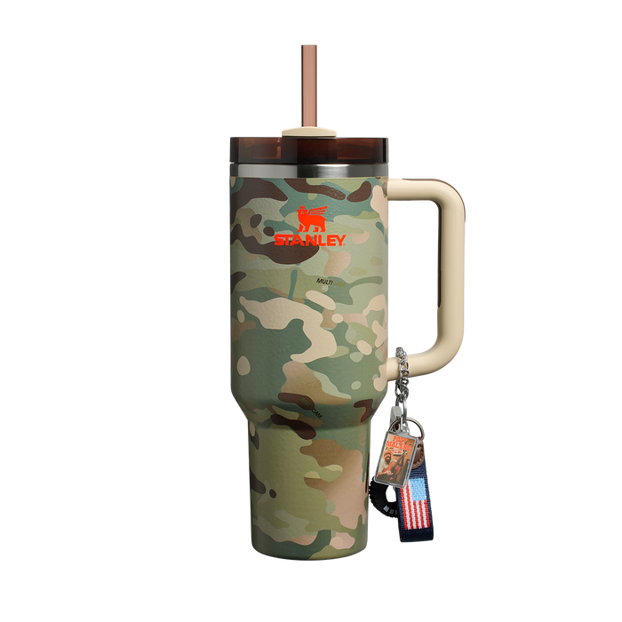 Mostra um copo térmico "Stanley Quencher" com estampa camuflada, alça bege e tampa com canudo. Ele tem o logotipo "Stanley" em vermelho e branco. Pendurado na alça, há um chaveiro com uma bandeira dos EUA e um pingente com a inscrição "Best Dad Ever" e uma imagem de um homem.