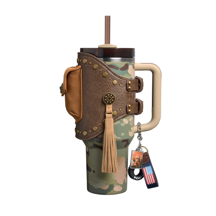 Um copo térmico camuflado com alça bege, tampa com canudo e faixa de couro marrom com tachas. Inclui bolso lateral, franja bege e chaveiro com bandeira dos EUA.